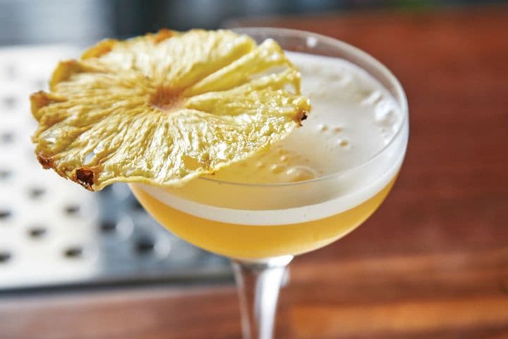 Foamy amber cocktail in a coupe glass topped with a large, thin dried pineapple garnish.